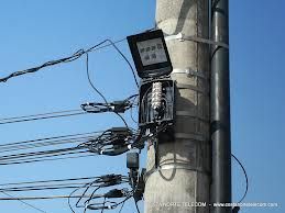 Poste com caixa de telecomunicações e cabos conectados, céu azul ao fundo.