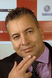 Man in suit with striped shirt and polka dot tie, posing with fingers near his chin.