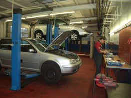 Autowerkstatt mit zwei Autos auf Hebebühnen, Mechaniker arbeitet an einer Werkbank.