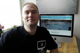 Persoon zit glimlachend voor een iMac op een tafel met een website op het scherm.