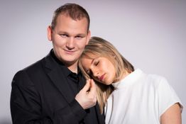 Ein Mann in Schwarz lächelt, während eine Frau im weißen Shirt ihren Kopf auf seine Schulter legt.