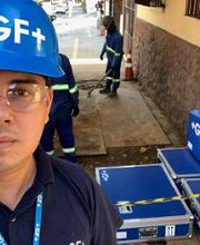 Homem com capacete azul "+GF+" em um local de trabalho ao ar livre, ao lado de caixas e cones.