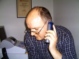 Mann mit Brille telefoniert, neben ihm ein Drucker und eine Urkunde an der Wand.