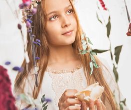 Niña con corona de flores, mirando al cielo, sosteniendo una concha entre flores colgantes.