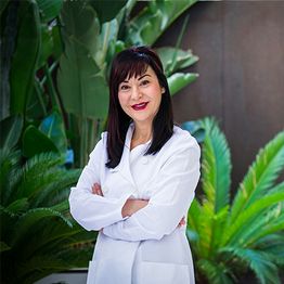 Doctora de pie con bata blanca, brazos cruzados, plantas verdes al fondo.