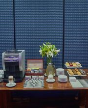 Mesa con cafetera, tazas, flores, galletas y refrescos junto a una pared azul texturizada.