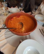 Cuenco de arroz caldoso en mesa con platos, cubiertos, y copas vacías. Personas al fondo.