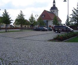 Kopfsteinpflaster-Parkplatz mit parkenden Autos, Bäumen und einer Kirche im Hintergrund.