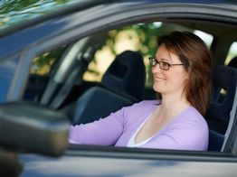 Frau mit Brille lächelt und fährt Auto, trägt lilafarbene Strickjacke.