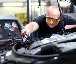 Mechaniker prüft Motor mit Werkzeug unter offener Motorhaube, trägt Schutzbrille, Werkstattausrüstung.