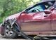 Damaged red car with broken windshield and bent metal against a backdrop of greenery.