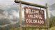 A wooden sign reads "Welcome to Colorful Colorado" against a mountain backdrop.