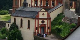 Kleine Kirche mit rotem Dekor, umgeben von Bäumen und Wiese, Menschen vor dem Eingang.