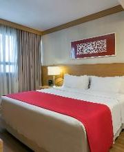 Quarto de hotel com cama de casal, colcha vermelha, luminárias e janela com cortina cinza.