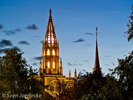 Kirchturm bei Dämmerung, beleuchtet, mit Baumumrissen gegen blauen Abendhimmel.