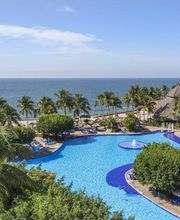 Complejo turístico con piscina, palmeras y vista al mar. Cielo despejado y edificios al fondo.