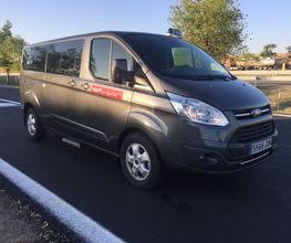 Furgoneta gris en una carretera asfaltada con árboles al fondo y un número de taxi en el parabrisas.