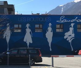 Wand mit stilisierten Modefiguren und Auto im Vordergrund, umgeben von Bergen, "Les Boutiques".