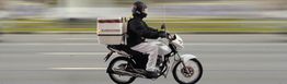 Motociclista de entrega dirigindo rápido em uma estrada com capacete e mochila de carga branca.