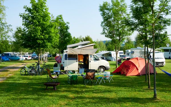 weißer Camper mit Markise auf einem Campingplatz im Sommer. Neben ihm steht ein orangenes Zelt auf einer grünen Wiese.