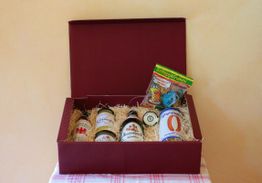 Rote Geschenkbox mit Flaschen und einer Süßigkeit auf Holzwolle, auf gestreiftem Tuch gelegen.