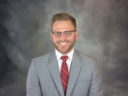 Smiling man in glasses and gray suit with red tie against a gray background.