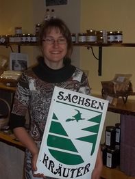 Frau hält ein Schild mit der Aufschrift "Sachsen Kräuter" in einem Laden mit Gläsern im Hintergrund.