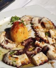 Plato de pulpo a la plancha con una papa asada en un plato blanco sobre mesa al aire libre.