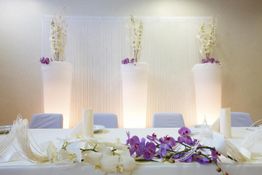 Weiße und lila Blumendeko auf einem Tisch vor drei beleuchteten Vasen mit weißen Blumen.