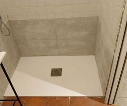 Ducha con plato blanco, azulejos grises, lavabo al lado y piso de cerámica marrón.