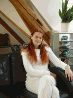 Eine Person mit roten Haaren sitzt in einem Raum auf einem Sofa, mit einer Pflanze im Hintergrund.