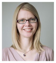 Eine lächelnde Frau mit Brille und blonden Haaren trägt ein rosa Oberteil vor grauem Hintergrund.