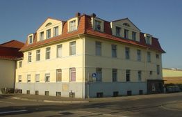 Gelbes, zweistöckiges Gebäude mit rotem Dach an einer Straßenecke unter blauem Himmel.