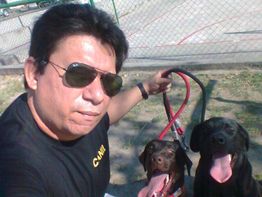 Homem com óculos escuros e camiseta preta segura dois cães pretos em uma área externa.