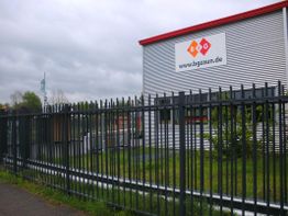Industriegebäude mit Firmenlogo hinter einem hohen Metallzaun an einem bewölkten Tag.