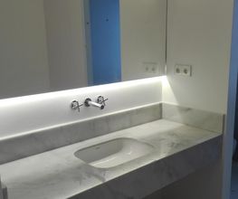Baño moderno con lavabo de mármol blanco, grifo en la pared e iluminación LED suave.