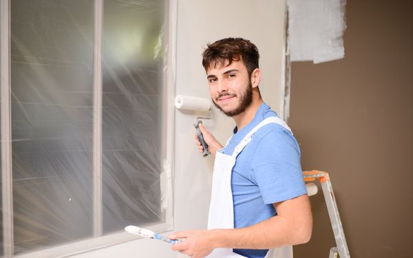 Ein Maler steht in einem Eck eines Wohnzimmers und grundiert eine Wand weiß. Das Fenster neben ihm ist zum Schutz vor Farbe abgeklebt.