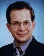 Ein Mann mit Brille und Anzug lächelt in die Kamera vor blauem Hintergrund.