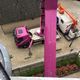 Zicht vanuit hoogte op roze en witte vrachtwagen met kraan naast gebouw.