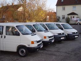 Fünf weiße Transporter parken nebeneinander auf einem Parkplatz vor einem Wohnhaus.