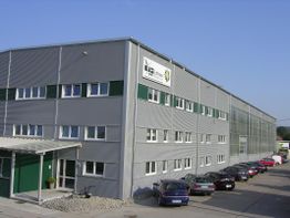 Graues Industrielager mit Fenstern, Autos parken davor unter blauem Himmel.
