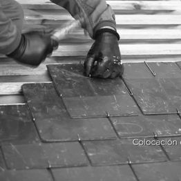 Persona colocando baldosas negras con guantes y herramientas en la mano. Texto: "Colocación".