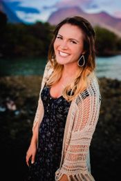 Woman smiling outdoors, wearing a patterned dress and crochet cardigan, with mountains in the background.