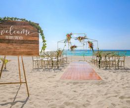 Boda en la playa con arco floral frente al mar y cartel de bienvenida sobre arena.