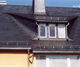Dach eines Hauses mit Schieferbedeckung und markanten Fenstern unter blauem Himmel.
