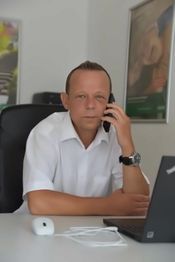 Ein Mann sitzt an einem Schreibtisch mit Laptop und Telefon, trägt ein weißes Hemd.
