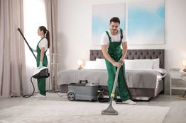Two cleaners in green overalls vacuuming a carpet in a modern bedroom setting.