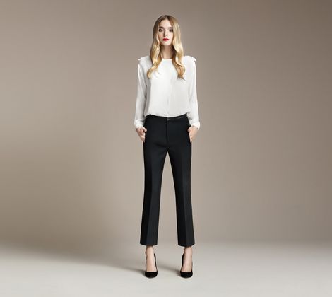 Una donna in piedi indossa camicia bianca e pantaloni scuri, sfondo neutro.