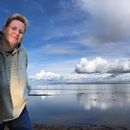 Eine Person lächelt am Ufer eines stillen Sees unter einem bewölkten blauen Himmel.