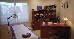 Sala de terapia con cama de masaje, escritorio de madera, estantería y lámpara de escritorio.